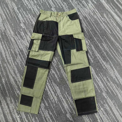 Front view of olive green and black color-block cargo pants laid flat on a gray patterned surface, showing belt loops, a button waistband, and flap thigh pockets.