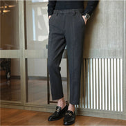 A person wearing dark gray textured trousers, a black long-sleeve top, and black loafers, standing indoors against a modern glass and wood-paneled wall.