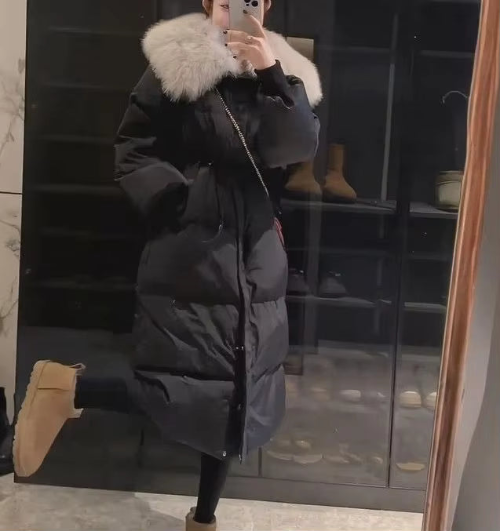 Model wearing a long black quilted coat with a large cream fur-look collar, drawstring waist and front zipper, paired with beige boots
