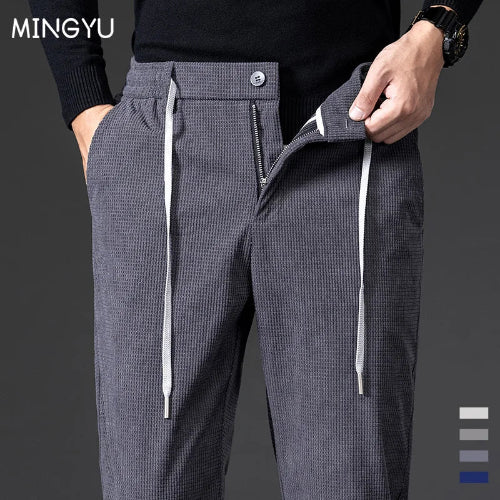 A close-up of a person wearing dark gray textured drawstring trousers with a front zipper and button closure, paired with a black sweater. The brand name MINGYU 