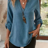 A woman wearing a blue long-sleeve linen shirt with rolled-up sleeves, a chest pocket, and a V-neckline, paired with black pants and layered necklaces.