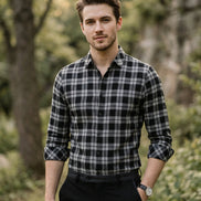 A man wearing a black and white plaid long-sleeve button-up shirt, paired with black trousers and a black belt, standing against a dark gray background.