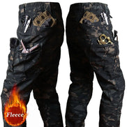 Two side views of dark camouflage cargo pants showing large thigh pockets and smaller pockets holding gloves, tools, and accessories against a white background.⠋