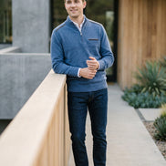 A man wearing a blue quarter-zip pullover sweater, dark blue jeans, and white sneakers stands on a modern outdoor walkway with concrete and wood architectural elements.