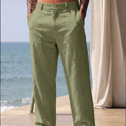 A man wearing light olive green linen trousers stands barefoot on a patio with the ocean and a cream curtain in the background.