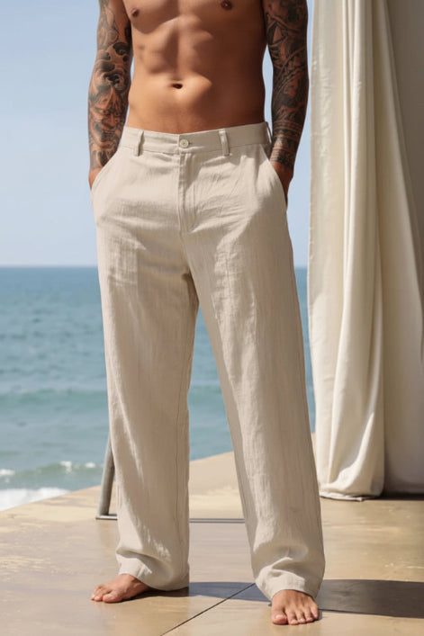 A man wearing light beige linen wide-leg trousers, standing barefoot on a stone deck with the ocean and a white curtain in the background.