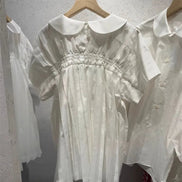 A white short-sleeve babydoll dress with a Peter Pan collar and smocked bodice, hanging on a wooden hanger on a clothing rack.