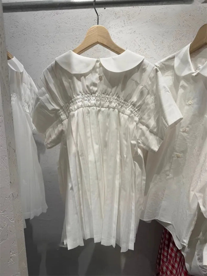 A white short-sleeve babydoll dress with a Peter Pan collar and smocked bodice, hanging on a wooden hanger on a clothing rack.