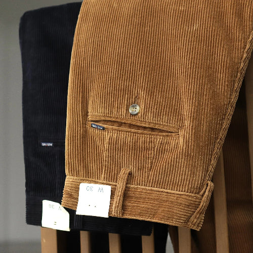 A close-up of brown corduroy trousers hanging on a display stand, showing the corduroy texture, a buttoned welt pocket, and a buttoned cuff. A black pair of the same trousers is partially visible behind them, along with white size tags.