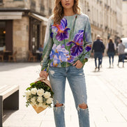 A woman wearing a long-sleeve crewneck sweatshirt with a watercolor-style print of purple, blue, and white irises on a muted green-gray background, paired with distressed blue jeans.