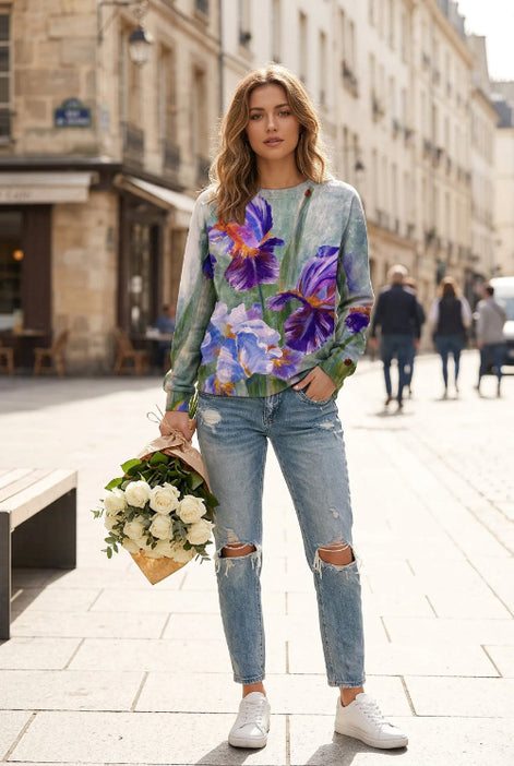 A woman wearing a long-sleeve crewneck sweatshirt with a watercolor-style print of purple, blue, and white irises on a muted green-gray background, paired with distressed blue jeans.