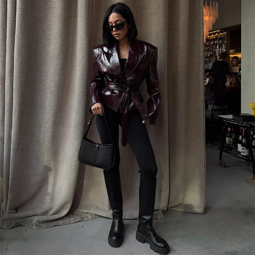 Woman wearing a glossy dark brown belted jacket with black fitted trousers, knee-high boots and sunglasses, holding a black handbag beside curtains