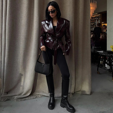 Woman wearing a glossy dark brown belted jacket with black fitted trousers, knee-high boots and sunglasses, holding a black handbag beside curtains