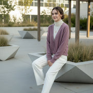 A person wearing a fuzzy mauve-pink V-neck cardigan over a cream turtleneck sweater, standing indoors near a window 