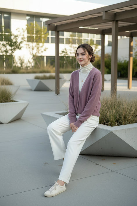 A person wearing a fuzzy mauve-pink V-neck cardigan over a cream turtleneck sweater, standing indoors near a window 