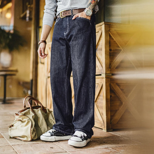 Person wearing dark blue jeans and black sneakers standing next to a green bag indoors.