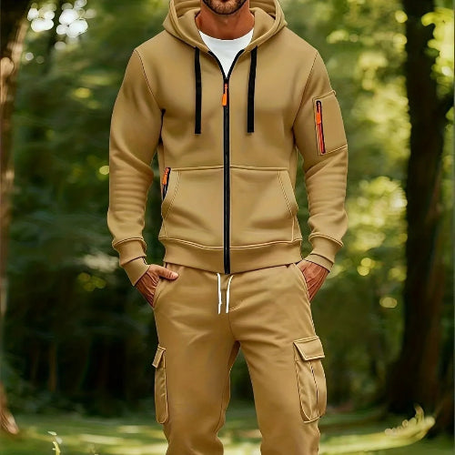 A man wearing a tan zip-up hoodie and matching cargo pants stands in a sunlit forest setting.