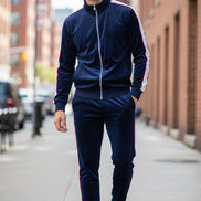A man modeling a matching navy blue velvet tracksuit with white piping details, featuring a zip-up jacket and matching pants, 