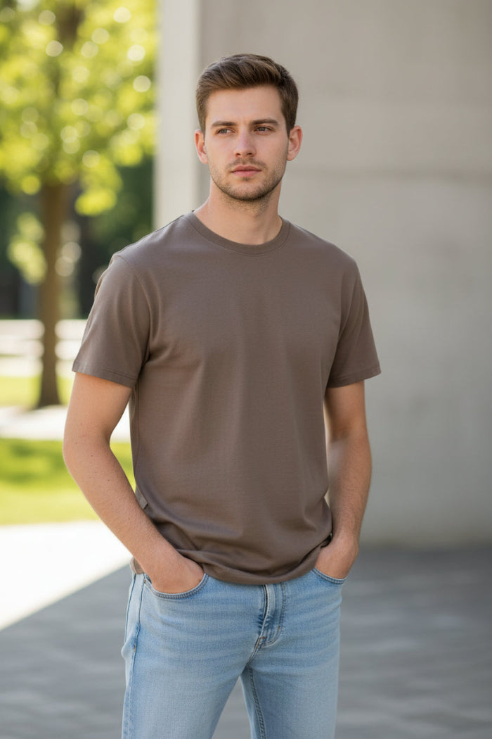 A man wearing a plain taupe-brown short-sleeve crewneck t-shirt and light blue jeans, shown from the chin down against a light gray background.