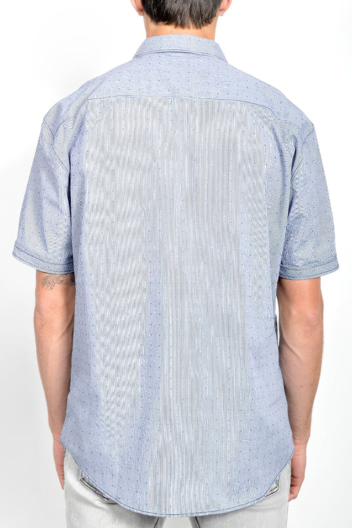 A man viewed from the back wearing a light blue short-sleeve button-down shirt with a subtle textured pattern, paired with light gray pants, against a plain white background.