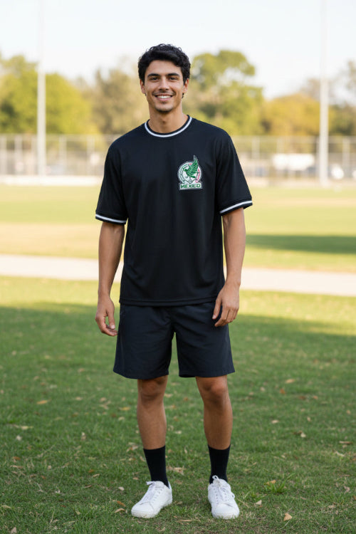 A man wearing a black short-sleeve t-shirt with white trim on the collar and cuffs, featuring a green and white Mexico national team crest 