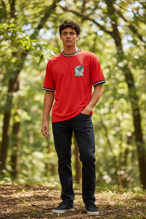 A man wearing a red short-sleeve t-shirt with black and white trim on the collar and cuffs, featuring the green and white Mexican national team crest on the left chest.
