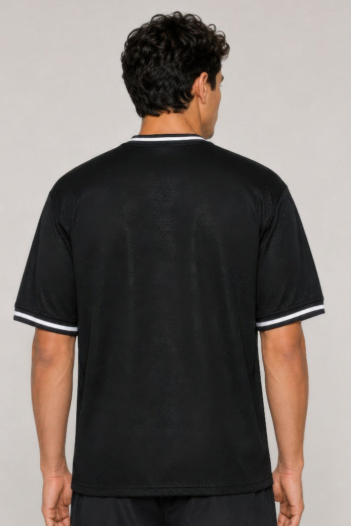 A man viewed from the back wearing a black short-sleeve t-shirt with white contrast trim on the collar and sleeve cuffs, against a plain light gray studio background.