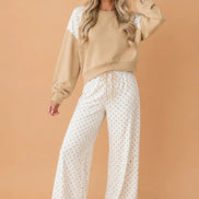 A woman modeling a two-piece lounge set consisting of a beige crewneck sweatshirt with white lace shoulder panels and white wide-leg pants 