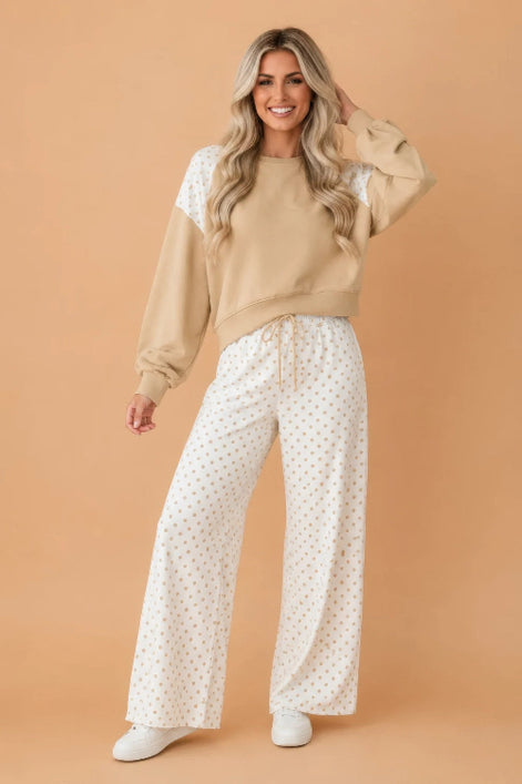 A woman modeling a two-piece lounge set consisting of a beige crewneck sweatshirt with white lace shoulder panels and white wide-leg pants 