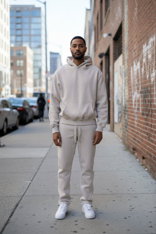 A man stands on a city sidewalk wearing a matching light beige hoodie and jogger sweatpants set with white sneakers