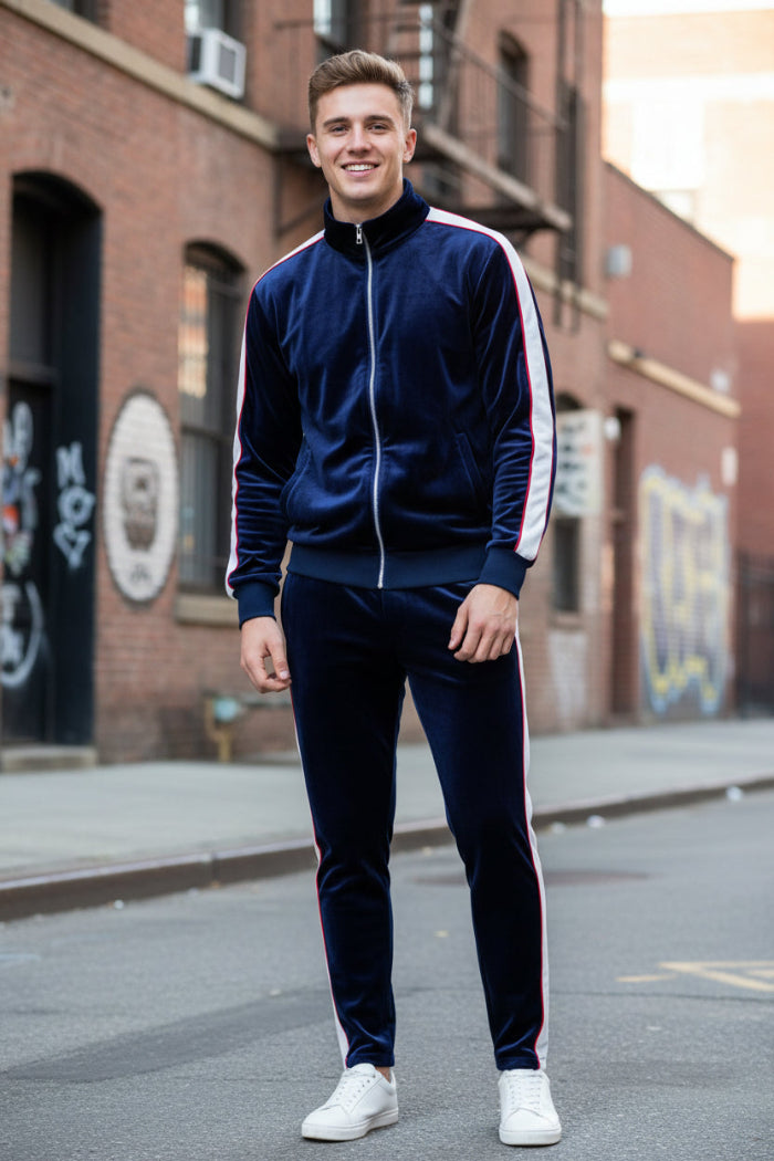 A man modeling a matching navy blue velvet tracksuit with white piping details, featuring a zip-up jacket and matching pants, paired with white sneakers  outside
