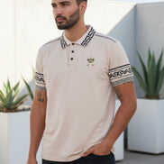 Man wearing a beige polo shirt with decorative patterns on sleeves and collar, standing against a white background.