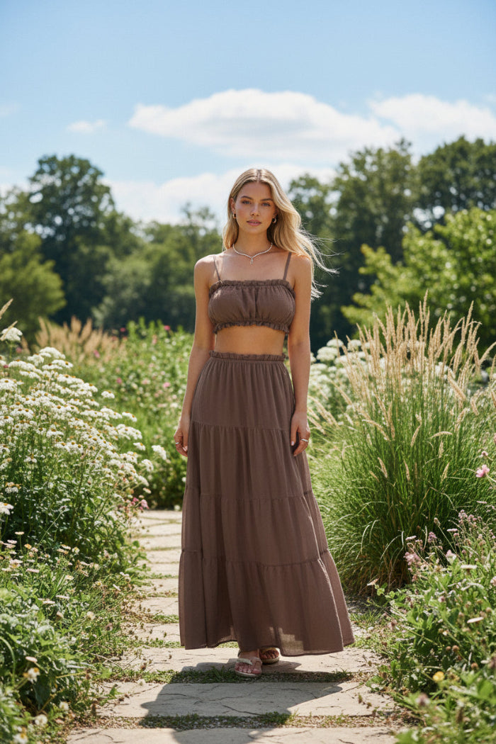 A woman wearing a two-piece brown set consisting of a cropped camisole top and a long tiered maxi skirt, standing in an indoor setting with warm-lit stone steps.