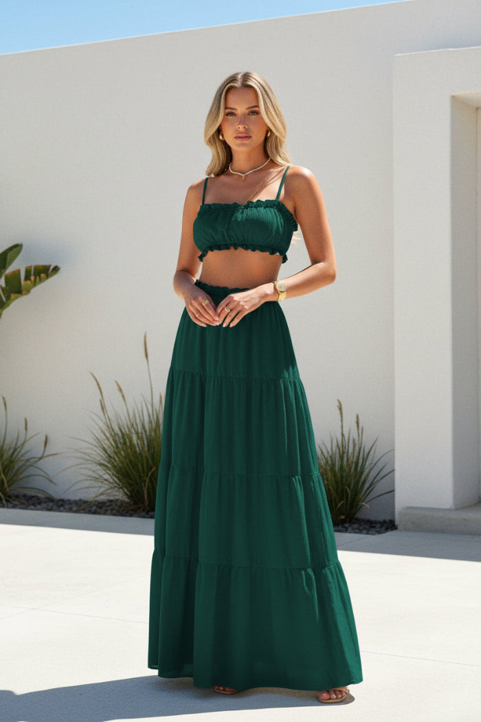 A woman modeling a dark green two-piece set consisting of a ruched crop top and a matching tiered maxi skirt, standing in a modern indoor setting with illuminated stone steps.