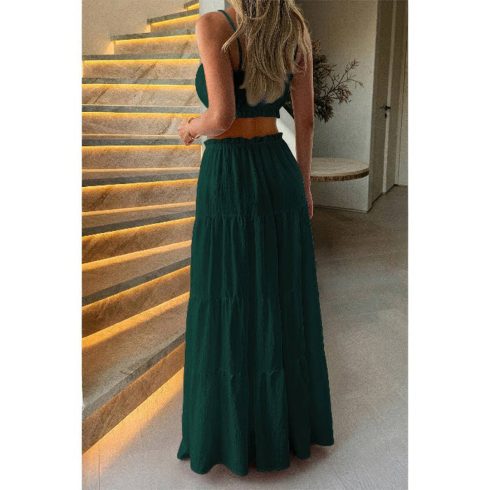 A woman wearing a dark green two-piece outfit consisting of a sleeveless crop top and a matching tiered maxi skirt, standing in front of an illuminated stone staircase.