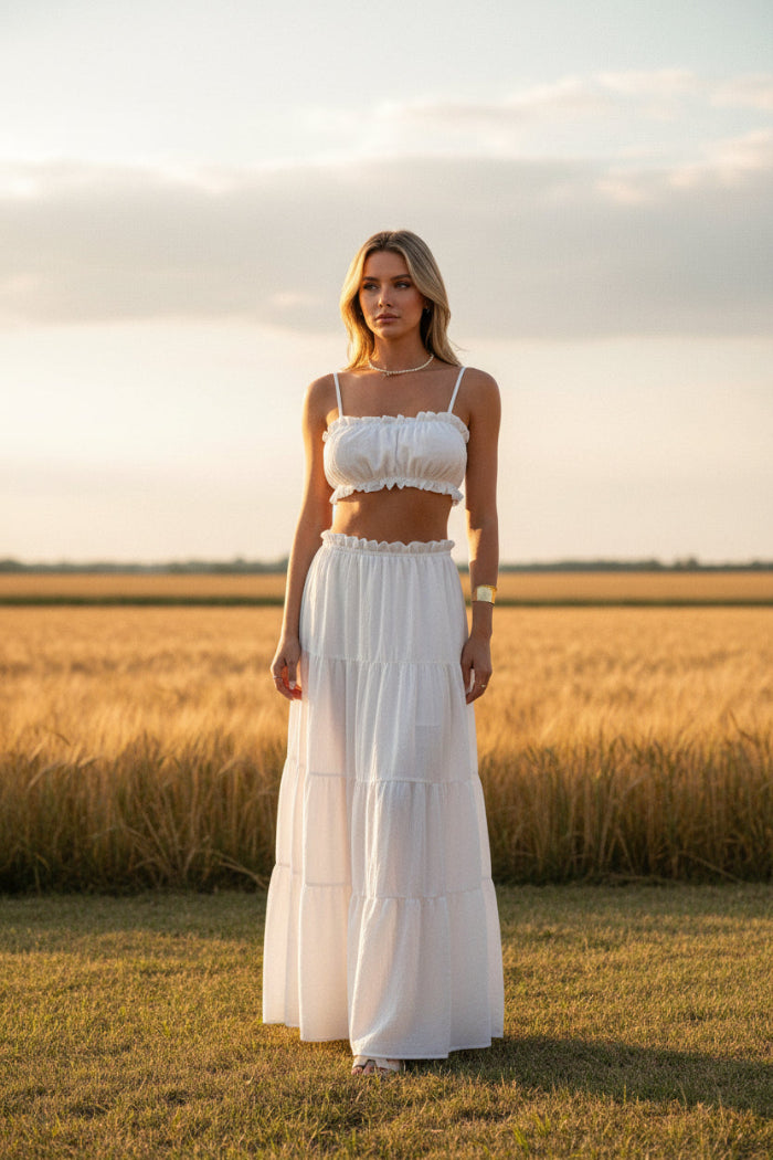 A woman modeling a white two-piece set consisting of a cropped spaghetti-strap top and a matching tiered maxi skirt, standing in a modern indoor hallway with warm-toned steps on the left.