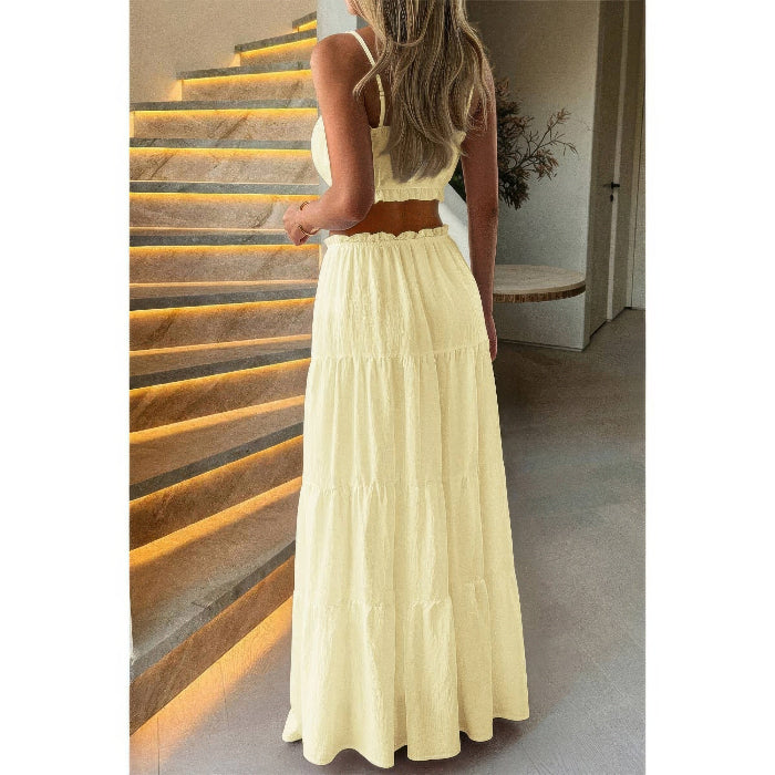 A woman wearing a light yellow two-piece dress with a sleeveless crop top and a long tiered skirt, standing in front of an illuminated staircase.