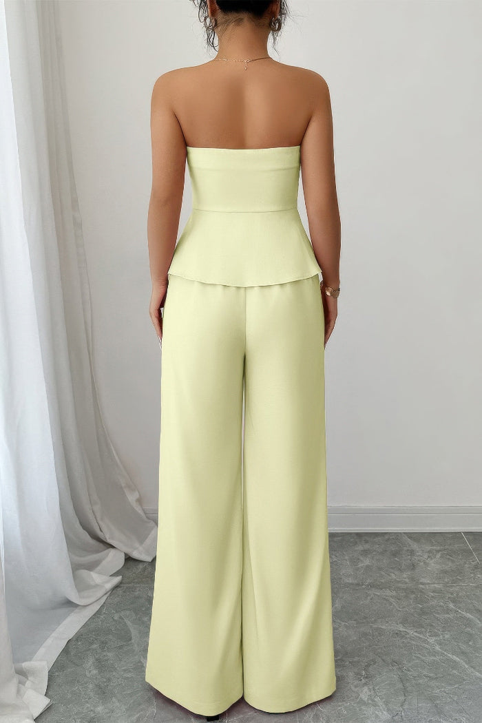 A woman shown from the back wearing a pale yellow strapless jumpsuit with a structured waist and wide legs, standing indoors against a light gray wall and white curtain.