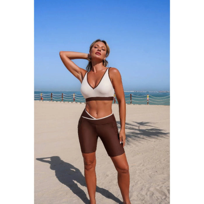 Woman in a white and brown sports bra and shorts standing on a beach with ocean view.