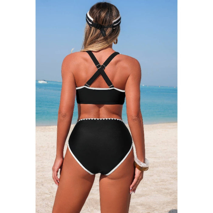 A woman stands on a sandy beach facing away from the camera, wearing a black high-waisted bikini with white trim and crisscross back straps.