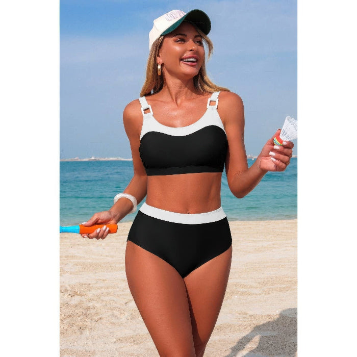 A woman modeling a two-piece bikini swimsuit on a sandy beach with the ocean and blue sky in the background. The swimsuit is black with white contrast trim, and she is also wearing a white cap and holding a small bottle and an orange toy.