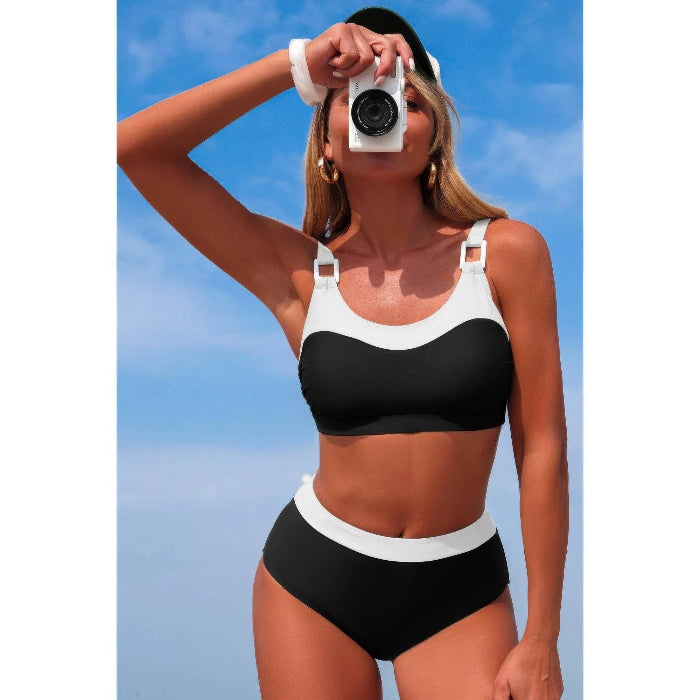 A woman modeling a black and white two-piece bikini with a white visor and white wristbands, holding a white camera against a blue sky background.