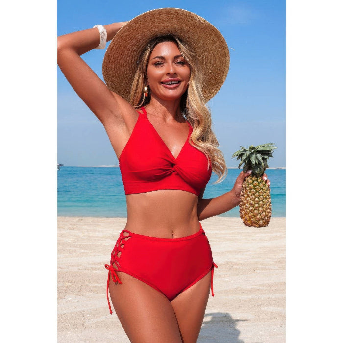 A woman on a beach wearing a bright red two-piece bikini and a large straw hat, holding a pineapple. The background shows blue ocean water and a sandy beach.