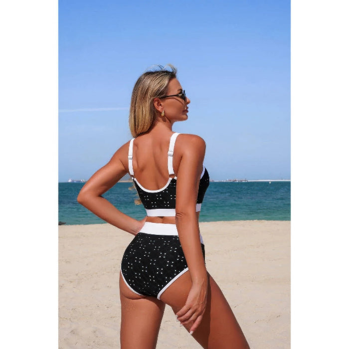 A woman wearing a black and white speckled two-piece swimsuit stands on a sandy beach with the ocean and blue sky in the background, photographed from behind.