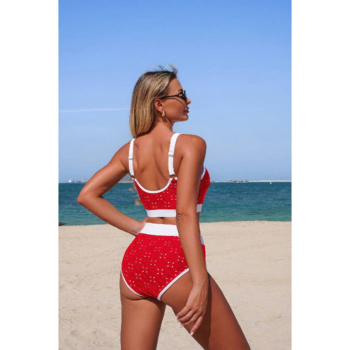 A woman wearing a red and white two-piece swimsuit with small white star patterns, standing on a sandy beach with the ocean and blue sky in the background.