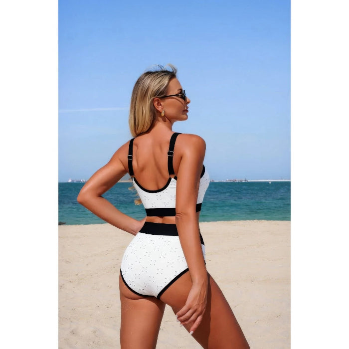 A woman wearing a white high-neck bikini with black trim and polka dots, standing on a sandy beach with the ocean and blue sky in the background.