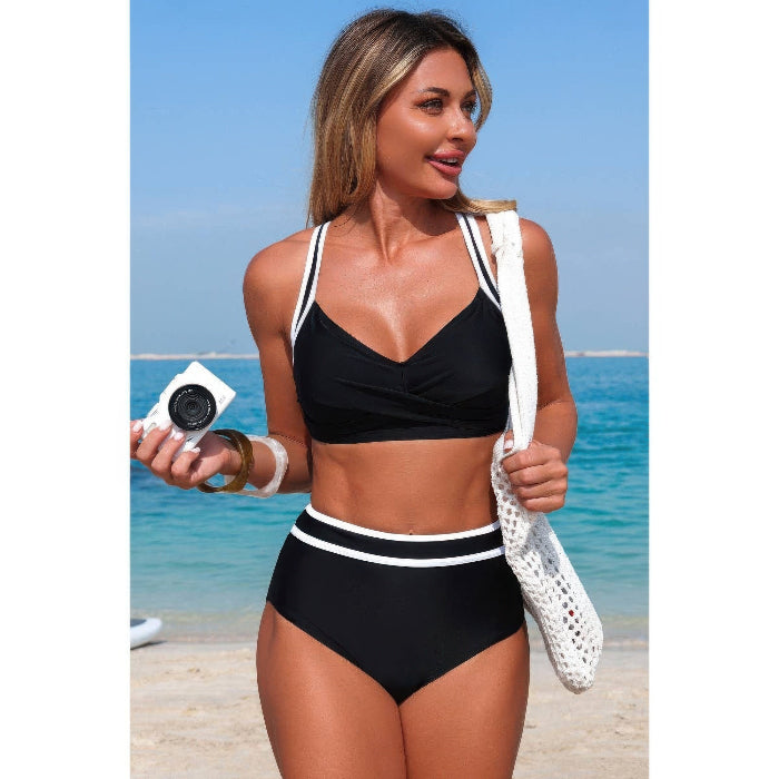 Woman in a black bikini with white straps standing on a beach holding a camera.
