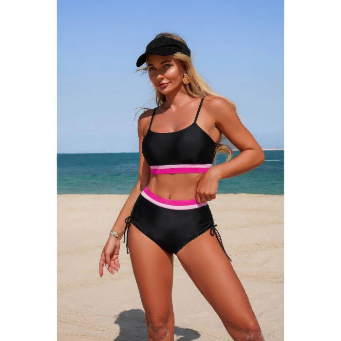 A woman modeling a two-piece black bikini with bright pink and white waistband stripes on a sandy beach with the ocean and blue sky in the background.