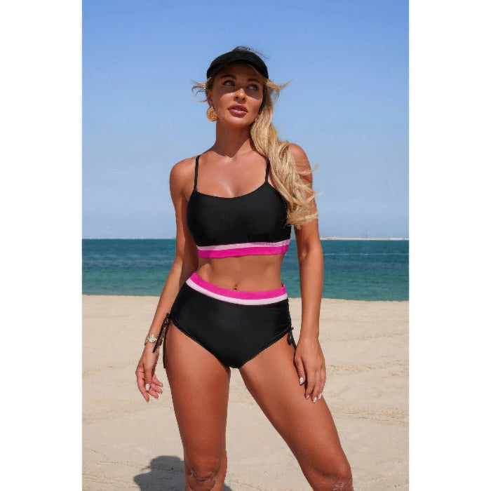 A woman modeling a two-piece black bikini with bright pink and white trim on a sandy beach with the ocean and blue sky in the background.