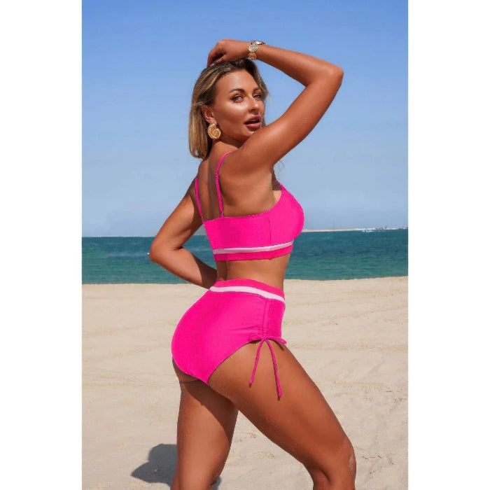 A woman posing on a sandy beach wearing a bright pink two-piece swimsuit with white stripes, with the ocean and blue sky in the background.